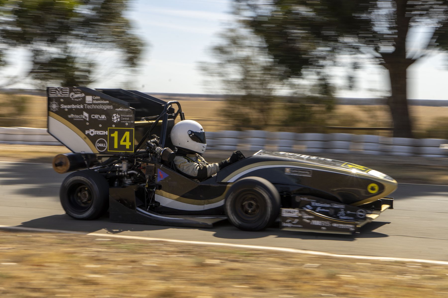 A black and gold Formula SAE car number 14 at speed on a track, driver in a white helmet